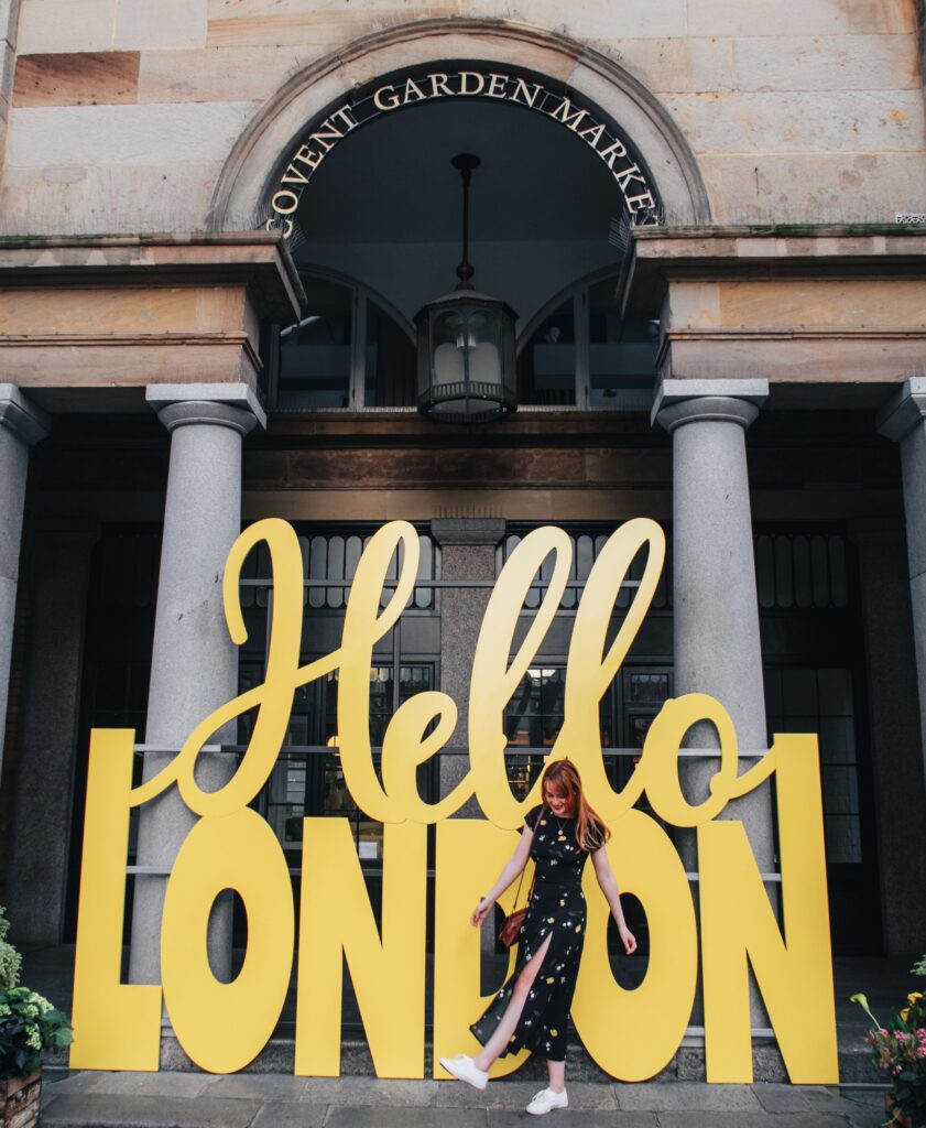 travel blogger stood infront of the hello London sign in Covent Market